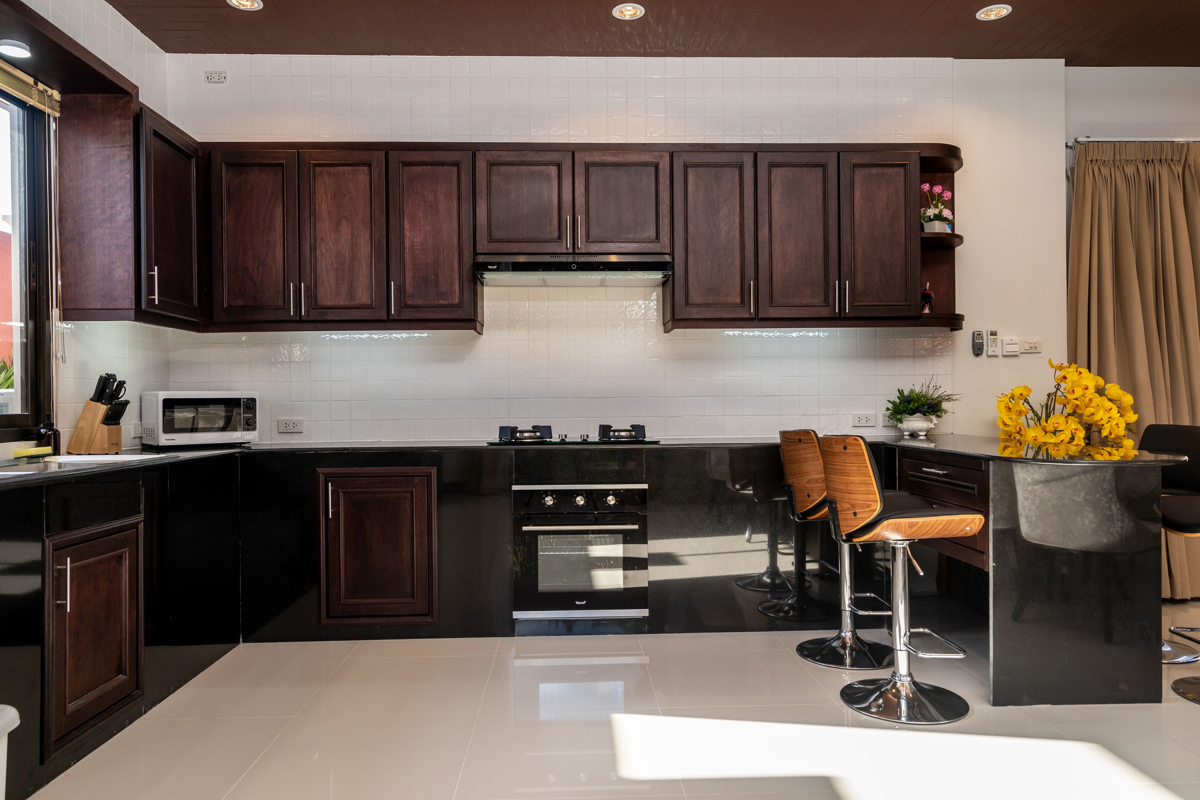 A modern kitchen with dark wood cabinets, a black countertop, a built-in oven, and a bar area with two stools.