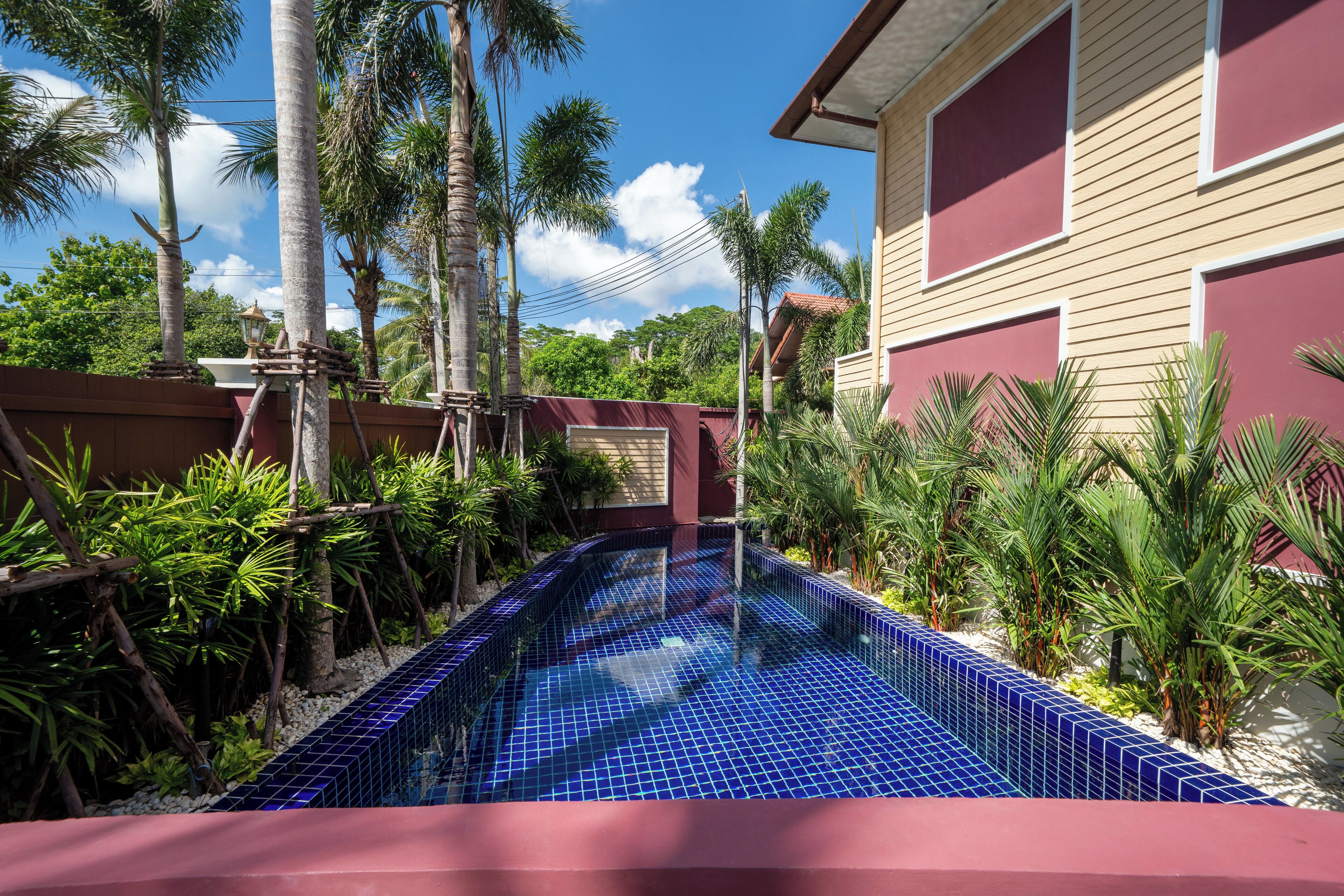 A swimming pool with blue tiles, surrounded by palm trees and plants, with a building in the background.