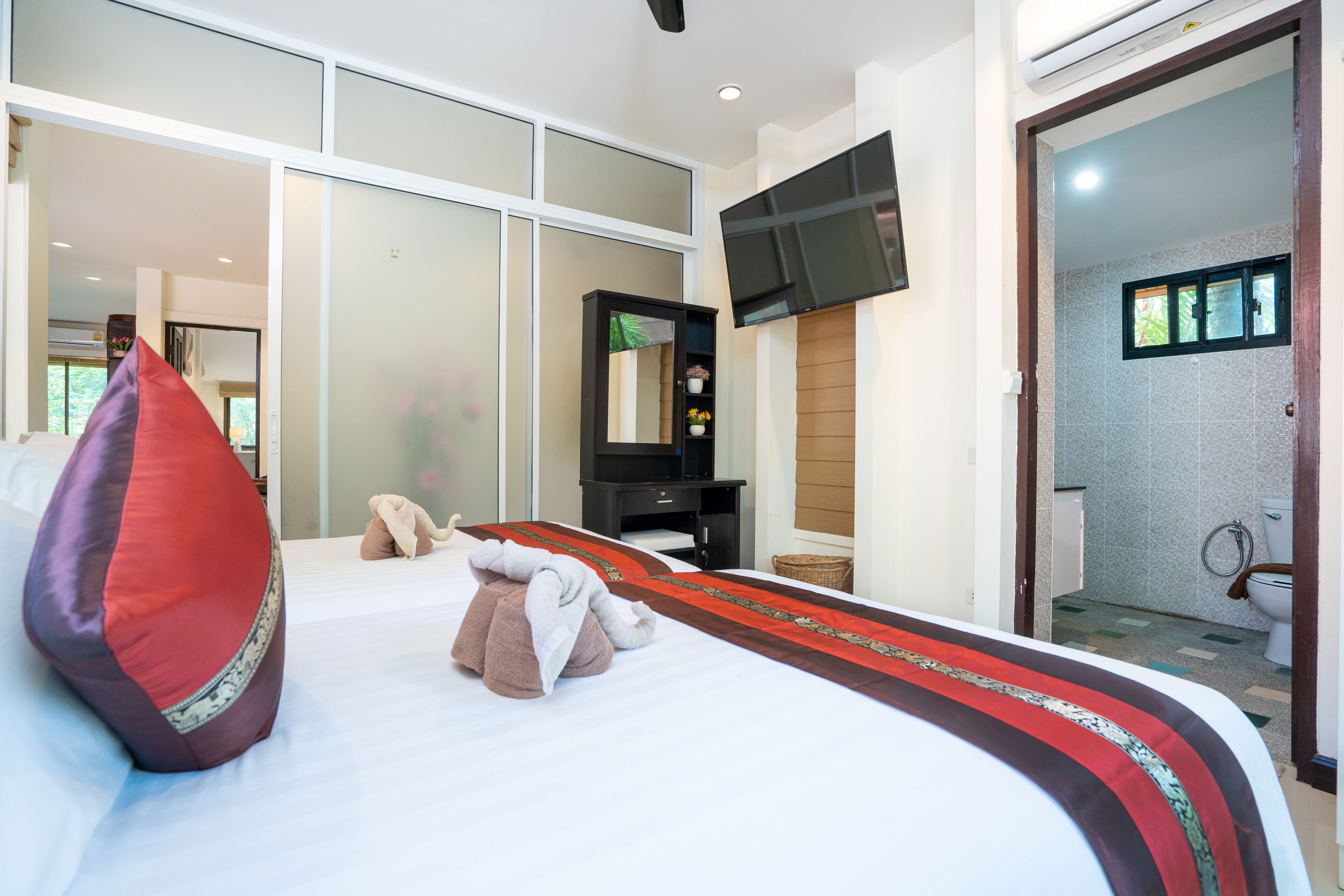 A hotel room with a large bed, a flat-screen TV mounted on the wall, a bathroom with a shower and toilet visible through an open door, and a small desk with a chair.