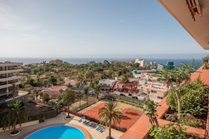 Zona de piscina con sillas de descanso y palmeras, con vistas a un pueblo costero y al mar.