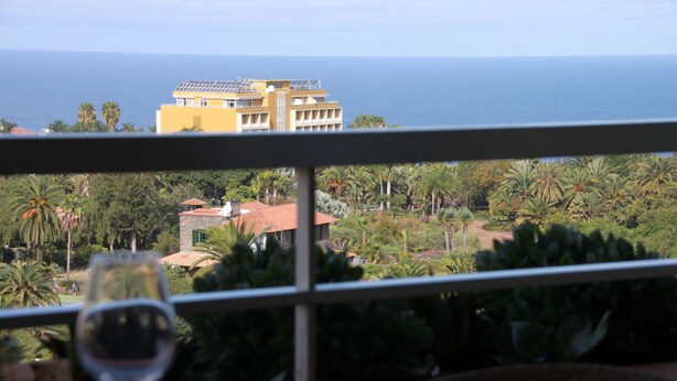 A balcony with a glass of wine, overlooking a yellow building, palm trees, and the ocean. - Apartment with fantastic panoramic views to the Teide and the sea, balcony facing west (Puerto de la Cruz)