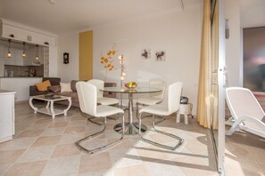 Una sala de estar moderna con una mesa de comedor de vidrio rodeada de sillas blancas, un sofá con cojines, una mesita auxiliar y un jarrón decorativo con flores.
