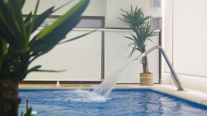 Une piscine agrémentée d’un élément décoratif aquatique, un pot de fleurs et un mur blanc avec une porte.