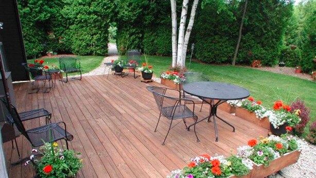 Una terraza de madera con mobiliario exterior, rodeada de macetas con flores y un área verde.
