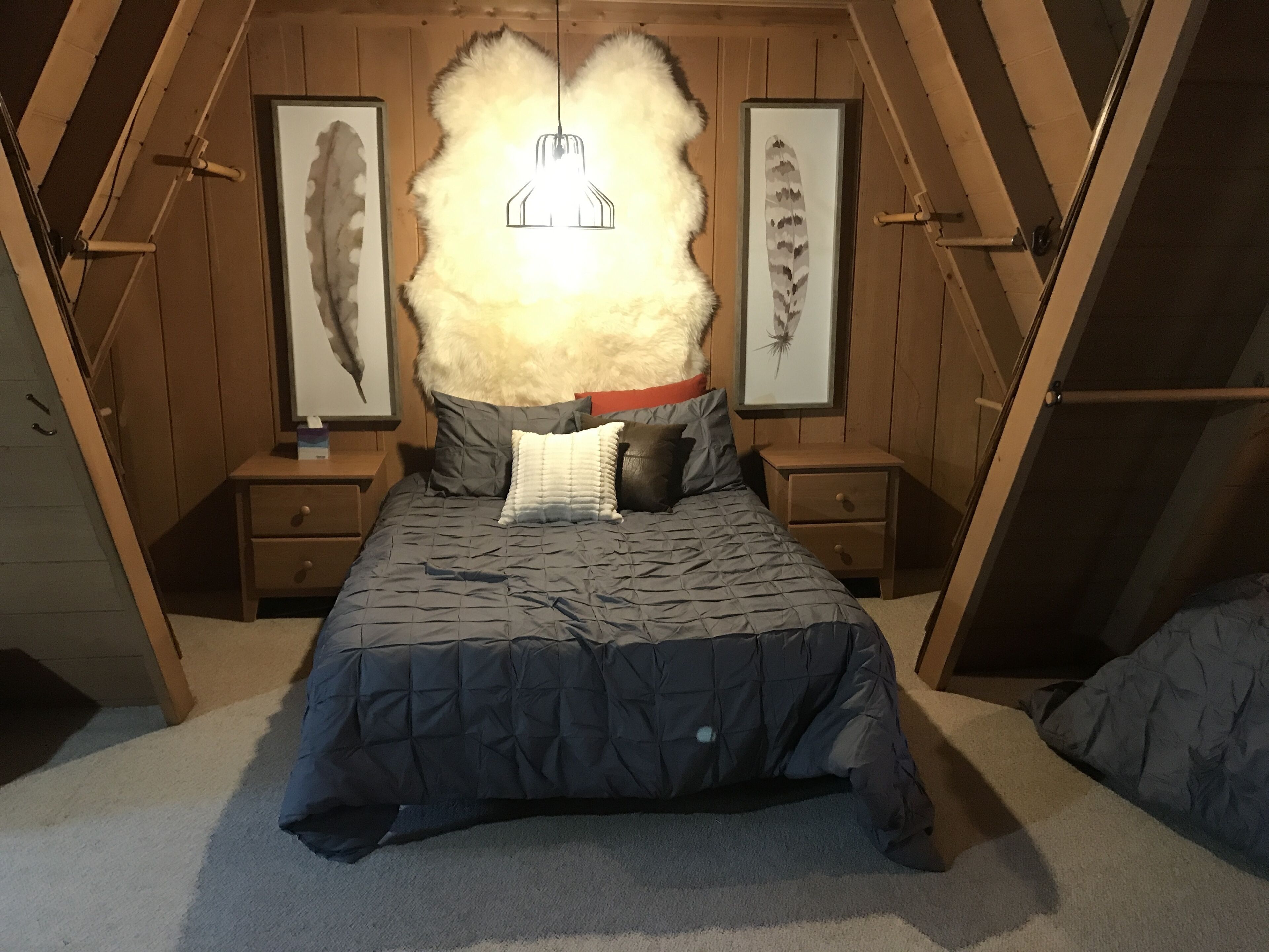 Une chambre mansardée avec un plafond en pente, un lit recouvert d’une courtepointe sombre, deux tables de chevet et des œuvres d’art représentant des plumes, encadrées, accrochées aux murs.