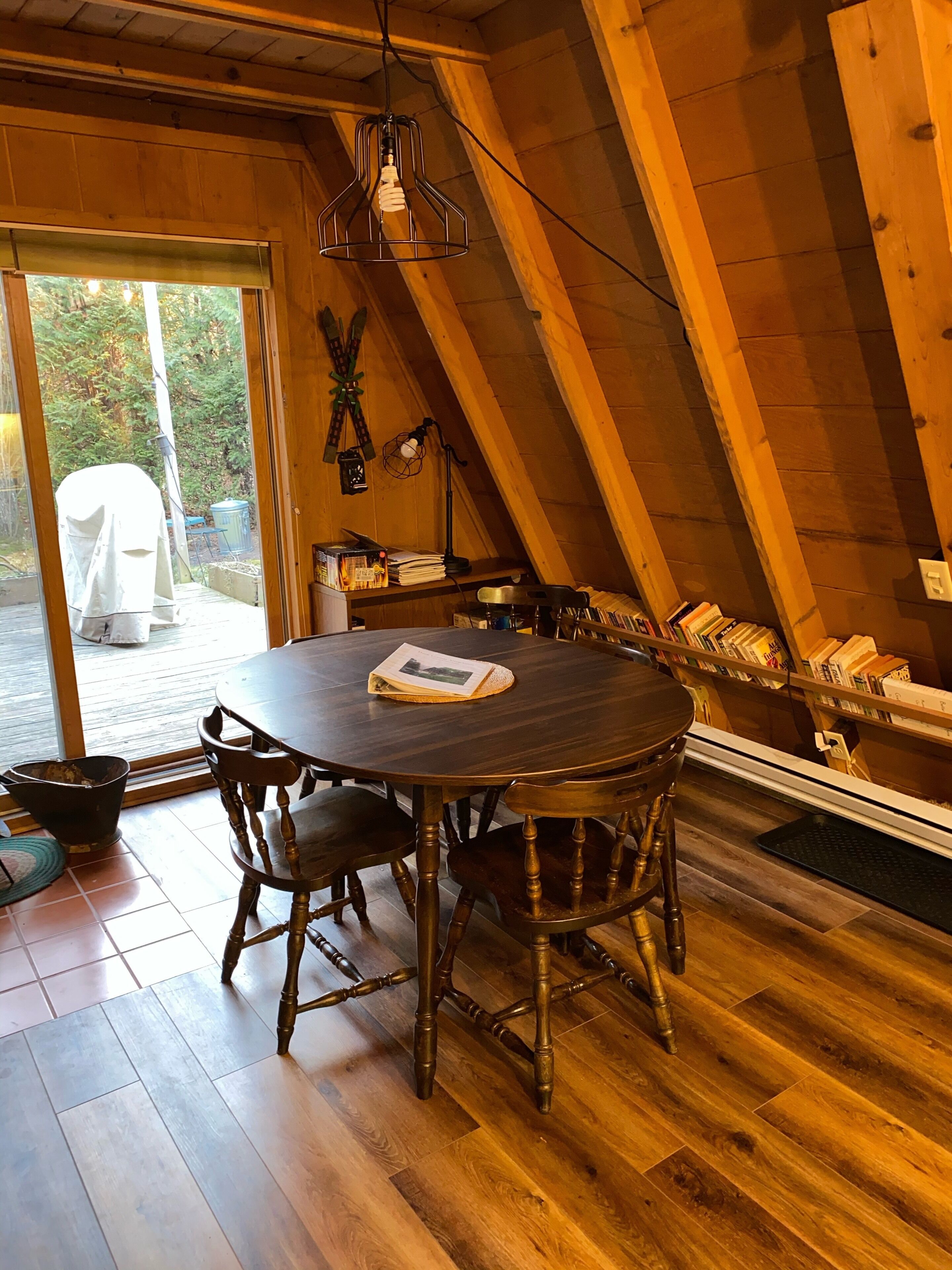 A cozy dining area with a wooden table and chairs, a hanging light fixture, and a bookshelf filled with books.