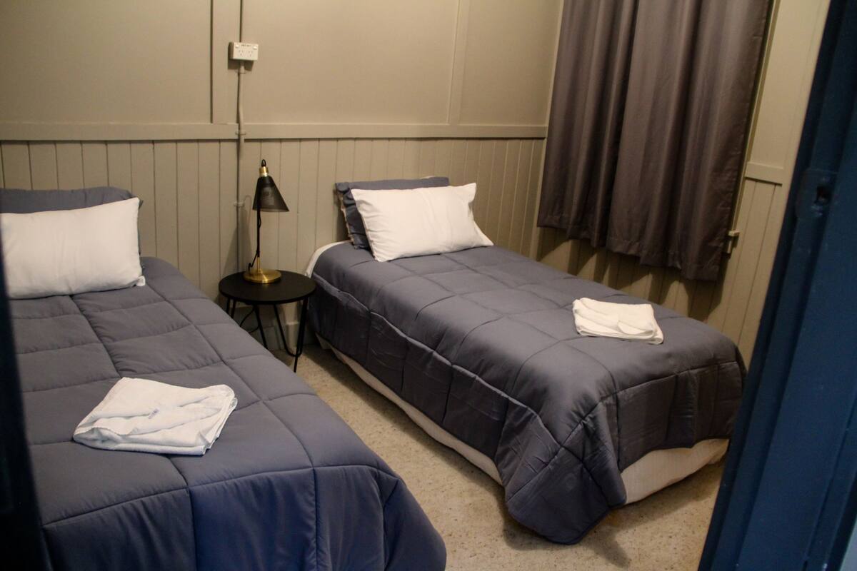 Deux lits simples avec une literie bleu foncé, une petite table ronde avec une lampe, et un mur sobre orné d’un interrupteur et d’un rideau.