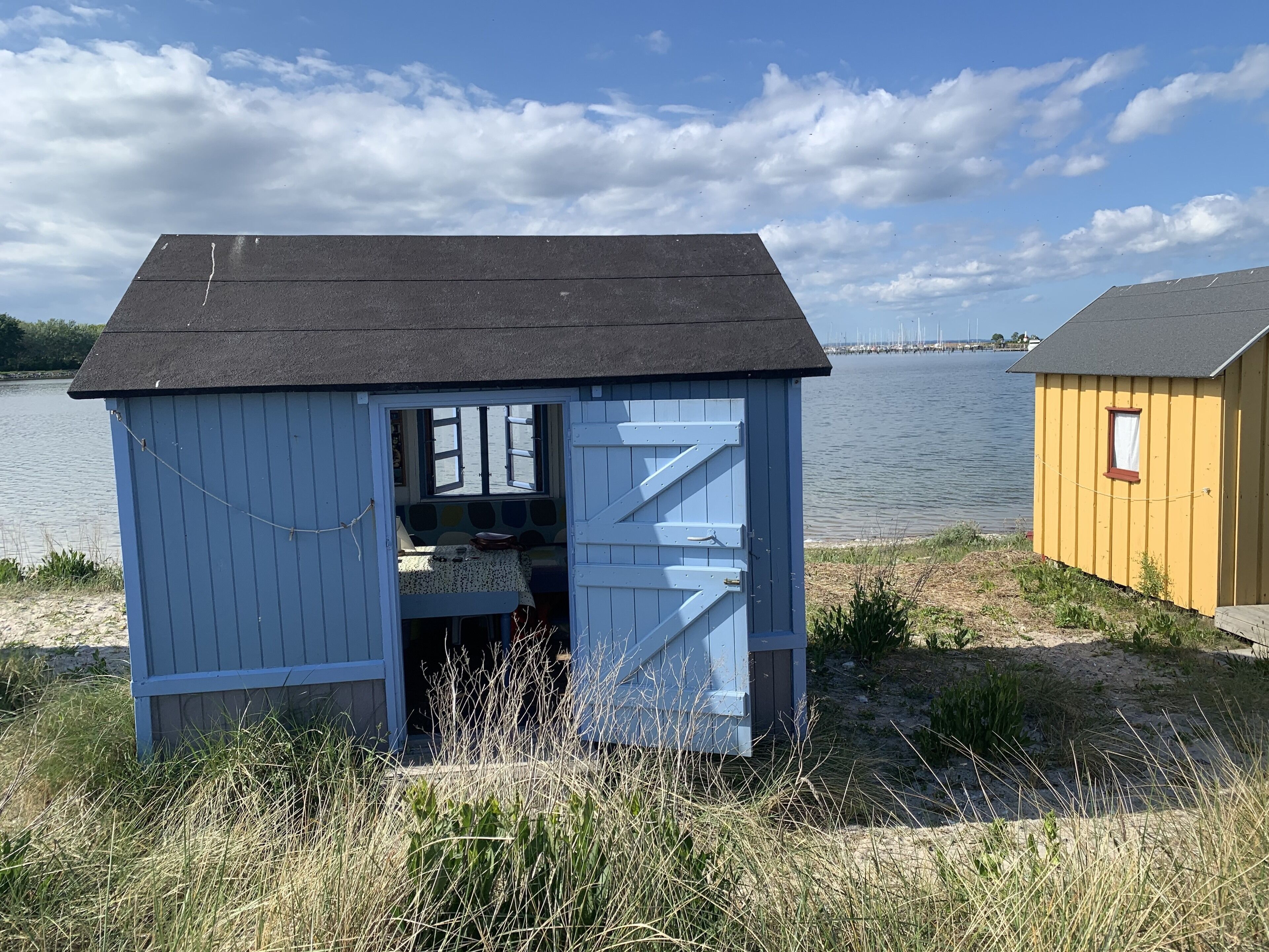 Een klein, blauw tuinhuisje met een geopende deur, waar een bed en een klein tafeltje in te zien zijn, gelegen aan een meer met een geel gebouw in de buurt.