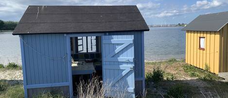 Een klein, blauw tuinhuisje met een geopende deur, waar een bed en een klein tafeltje in te zien zijn, gelegen aan een meer met een geel gebouw in de buurt.