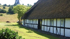 Ein traditionelles Fachwerkhäuschen mit Strohdach, gelegen in ländlicher Umgebung mit einer Windmühle im Hintergrund.