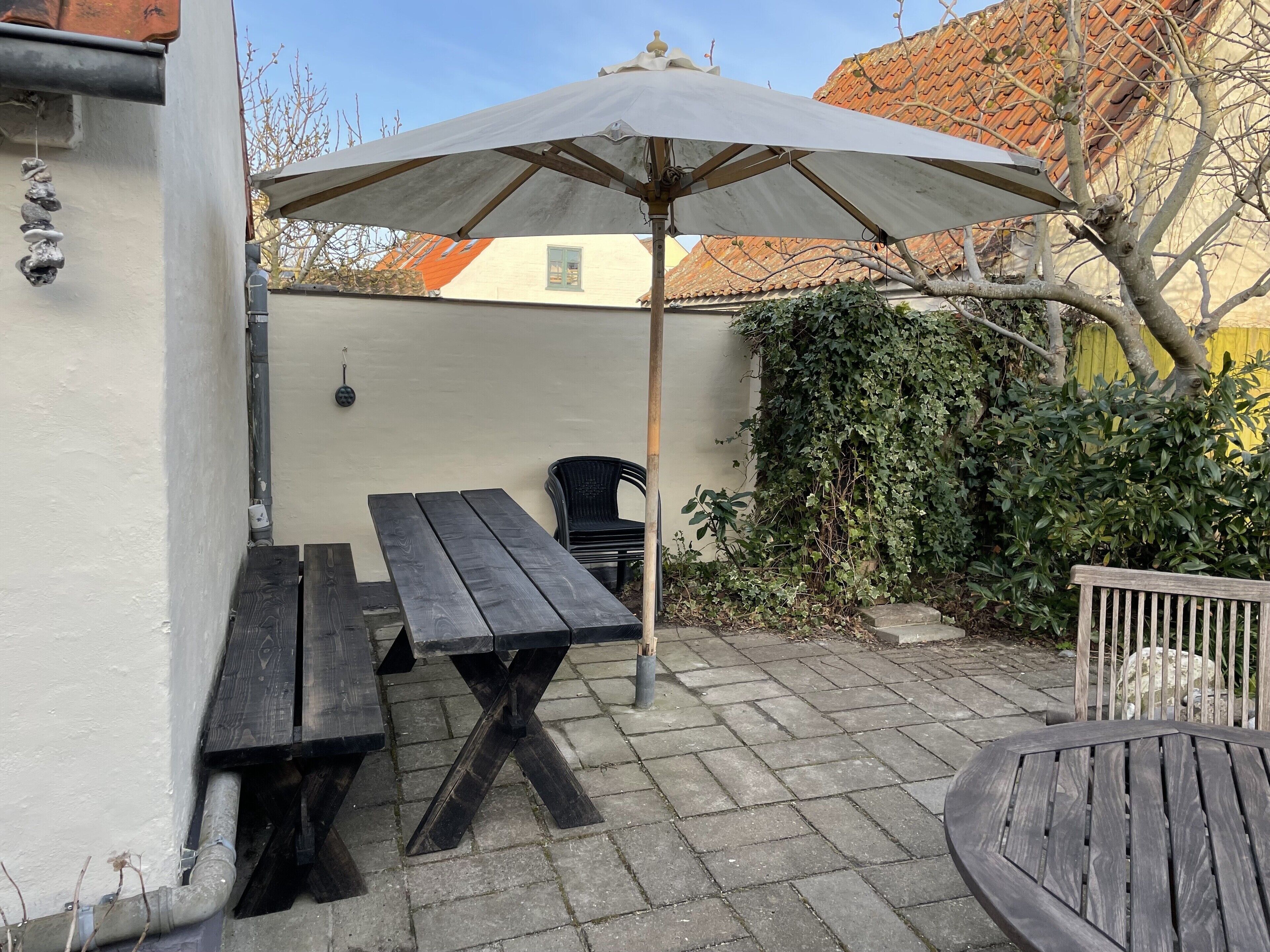 An outdoor patio with a wooden table and benches, a large umbrella, and a yellow wall in the background.