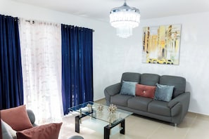 Un salon avec une table basse en verre, un canapé gris agrémenté de coussins, un lustre et un tableau représentant un paysage urbain accroché au mur.