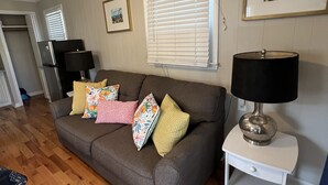 A living room with a grey sofa, wooden flooring, a white side table with a lamp, a framed picture on the wall, and a window with blinds. - Prime Location 1 Bedroom 1 Bath! (Carolina Beach)