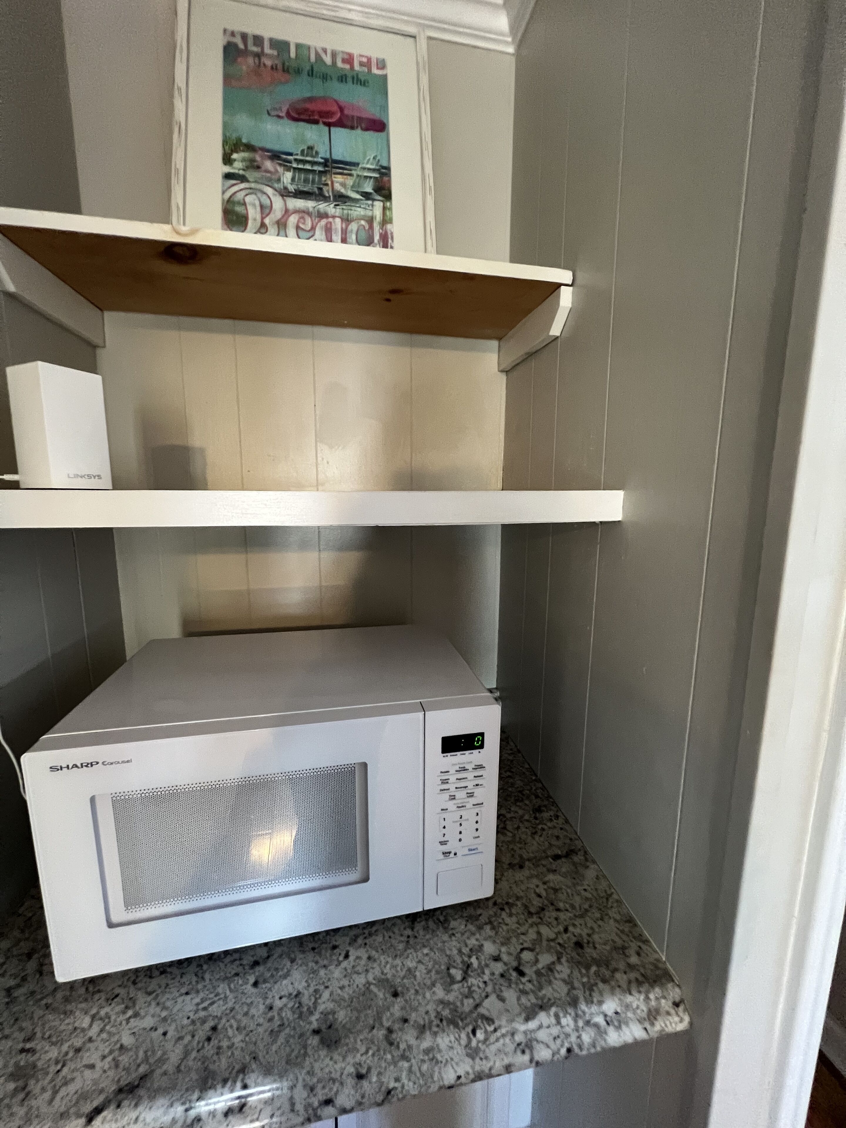 A kitchen with a microwave on a granite countertop, a wooden shelf above it, and a framed picture on the wall.