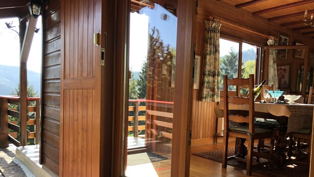 Uma cabana de madeira com uma porta de correr de vidro que dá para uma varanda. No interior, há uma mesa de jantar com cadeiras e um lustre.