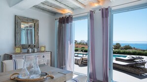 A room with a large window, a dining table with glassware, a mirror, and a view of a pool and landscape. - Aelia Luxury Villa with Sea view and swimming Pool (Paros)