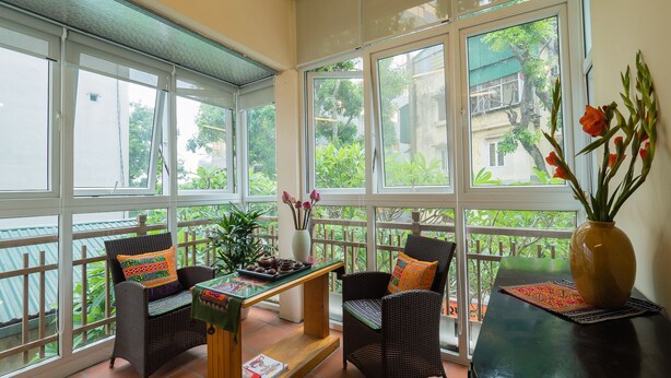 A sunlit room with large windows, wicker furniture, a small table with a vase of flowers, and a view of greenery outside. - Hanoi Balcony Homestay (Hanoi)