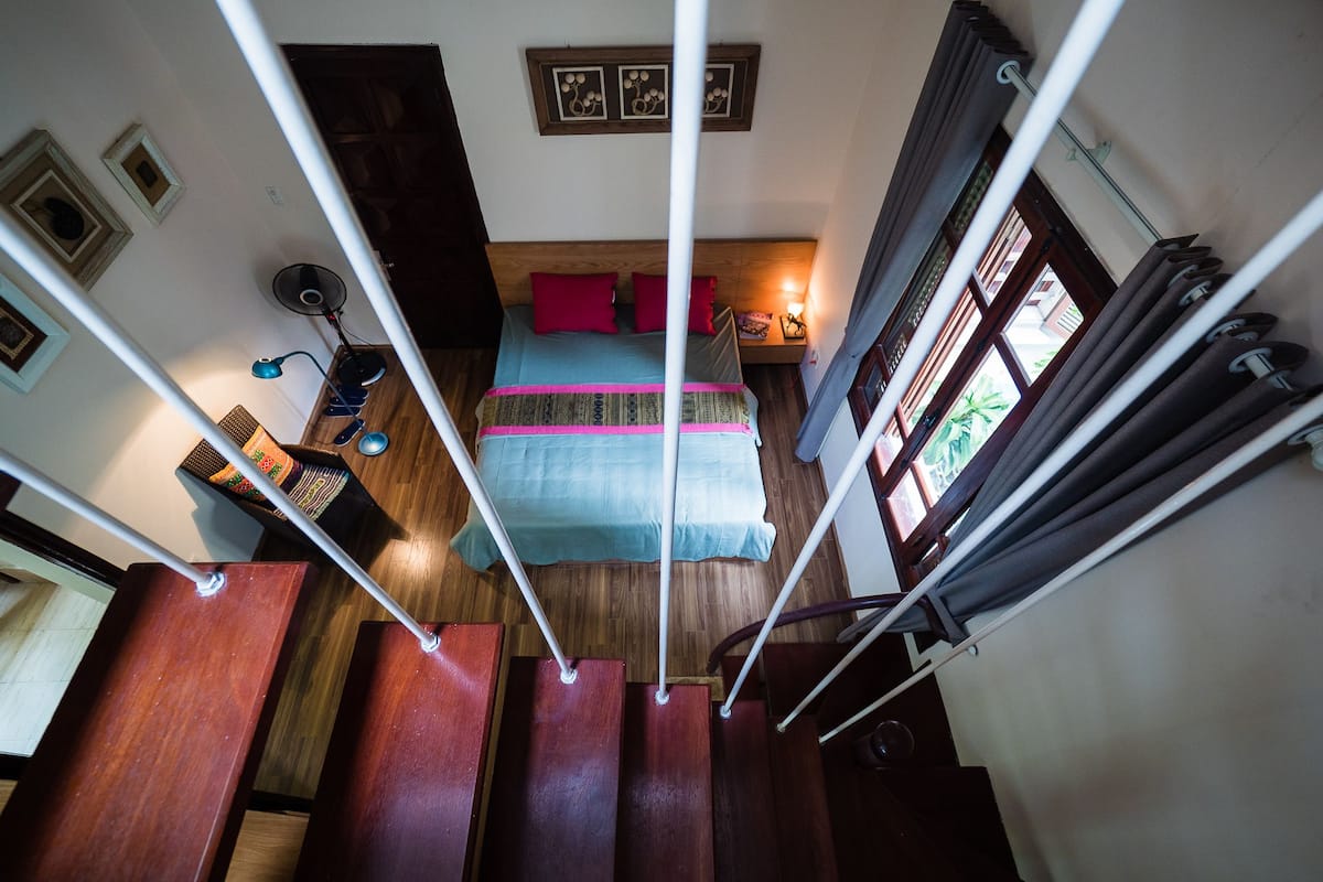Una escalera conduce a un dormitorio con cama, mesita de noche y ventana con cortinas.