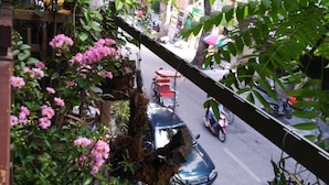 A street view with a parked car, a motorcycle, and a bicycle. There are trees and plants in the foreground. - Hanoi Balcony Homestay (Hanoi)