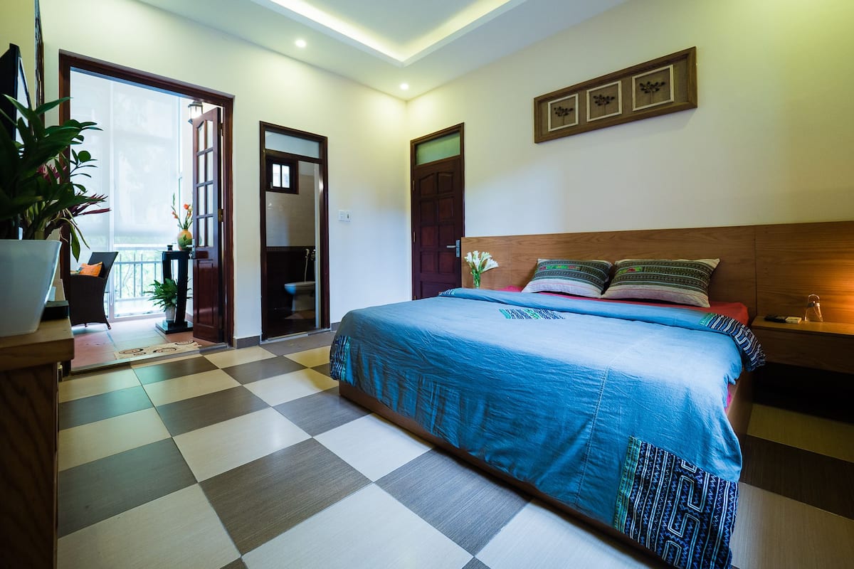 Un dormitorio con piso a cuadros, una cama con ropa de cama estampada, una mesita de noche, una lámpara de noche, una planta en maceta y una puerta que da a otra habitación.