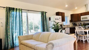 Ein Wohnzimmer mit Sofa, einem Esstisch mit Stühlen, einer Küchenzeile inklusive Schränken und Geräten sowie einem großen Fenster mit Vorhängen.