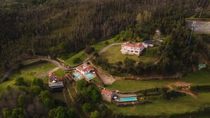 An aerial view of a rural property with multiple buildings, including a main house with a red roof, a smaller white building, and several structures with solar panels. There are multiple swimming pools and a winding road connecting the buildings.