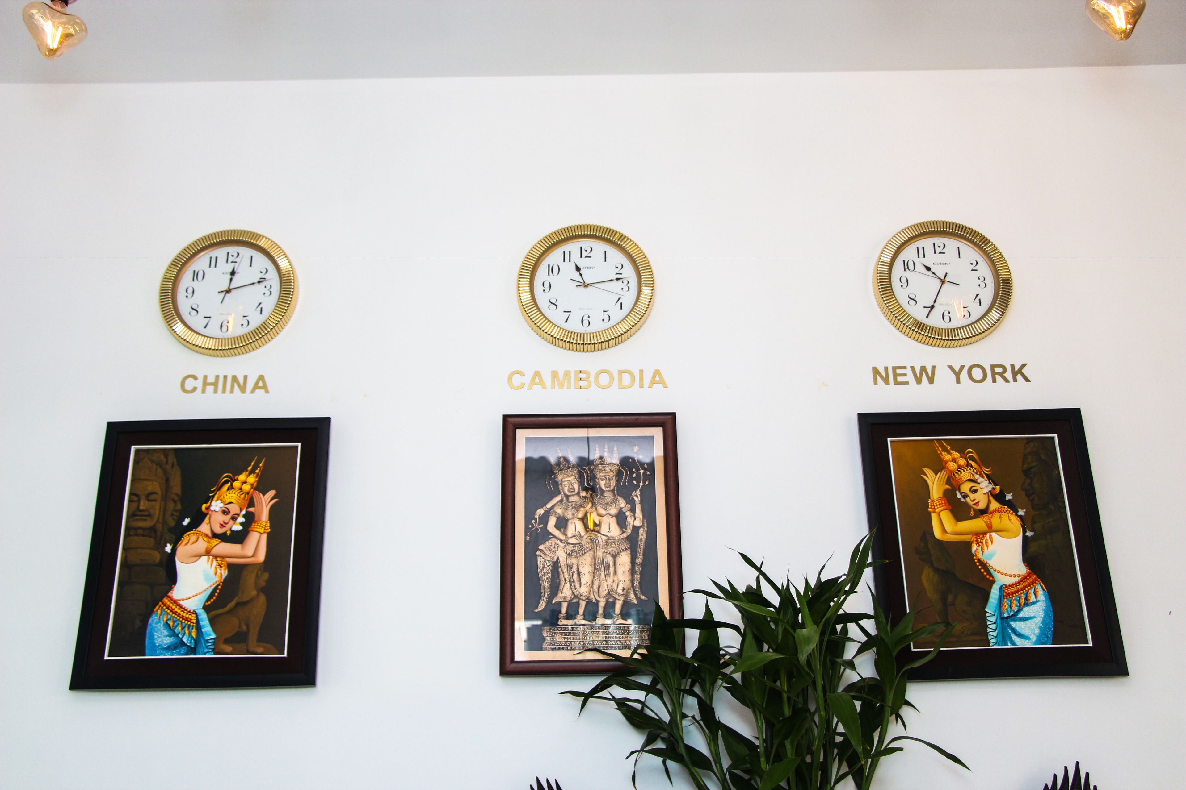 Three clocks labeled "CHINA," "CAMBODIA," and "NEW YORK" on a wall, each with a framed picture below.