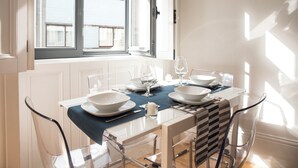 Mesa de comedor para cuatro personas con sillas de vidrio transparente, mantel blanco y vistas al exterior a través de una ventana.