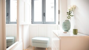Una habitación con ventana, un pequeño banco, un florero con flores y una taza verde sobre una superficie blanca.