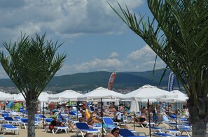 A beach with rows of blue lounge chairs and white umbrellas, a palm tree, and a flag in the background.