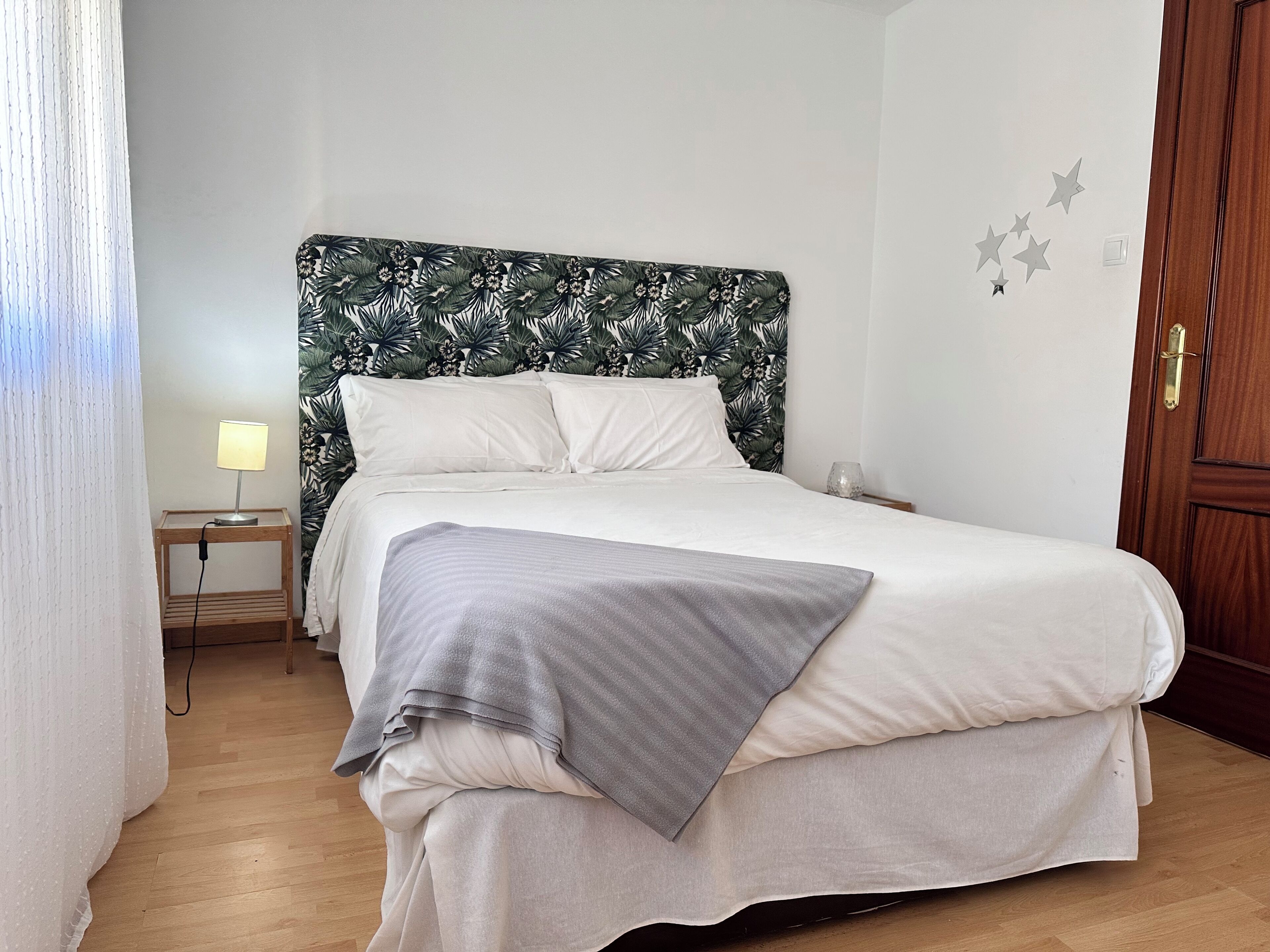 Ein ordentlich bezogenes Bett mit grauer Decke, ein Nachttisch mit Lampe und ein Beistelltischchen mit einem Glas.