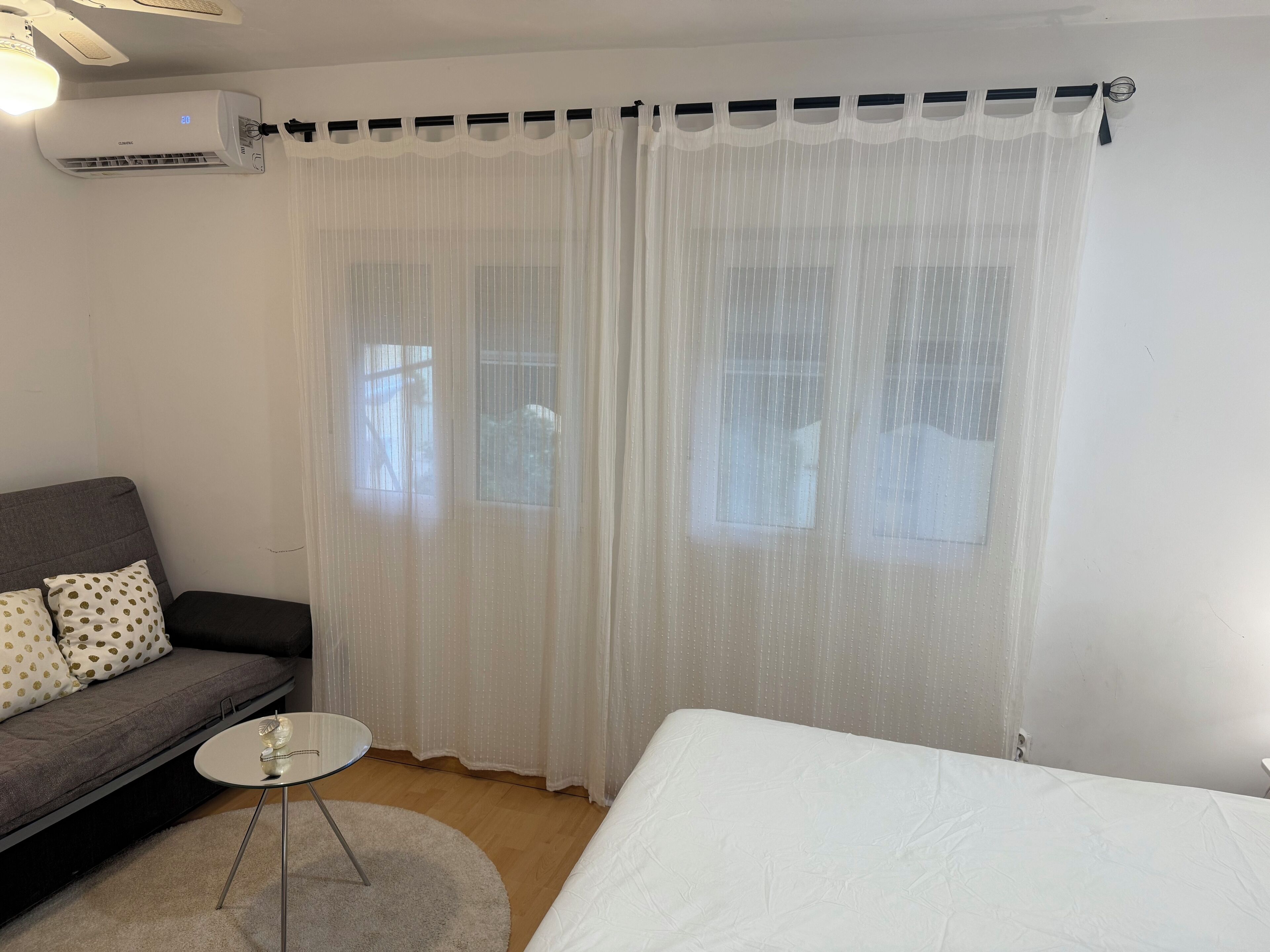 Una habitación con cama, un sofá con cojines estampados, una mesita redonda y cortinas de encaje.