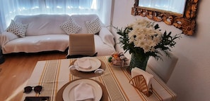 Uma área de refeições com uma mesa posta para dois, um sofá e um vaso decorativo com flores.