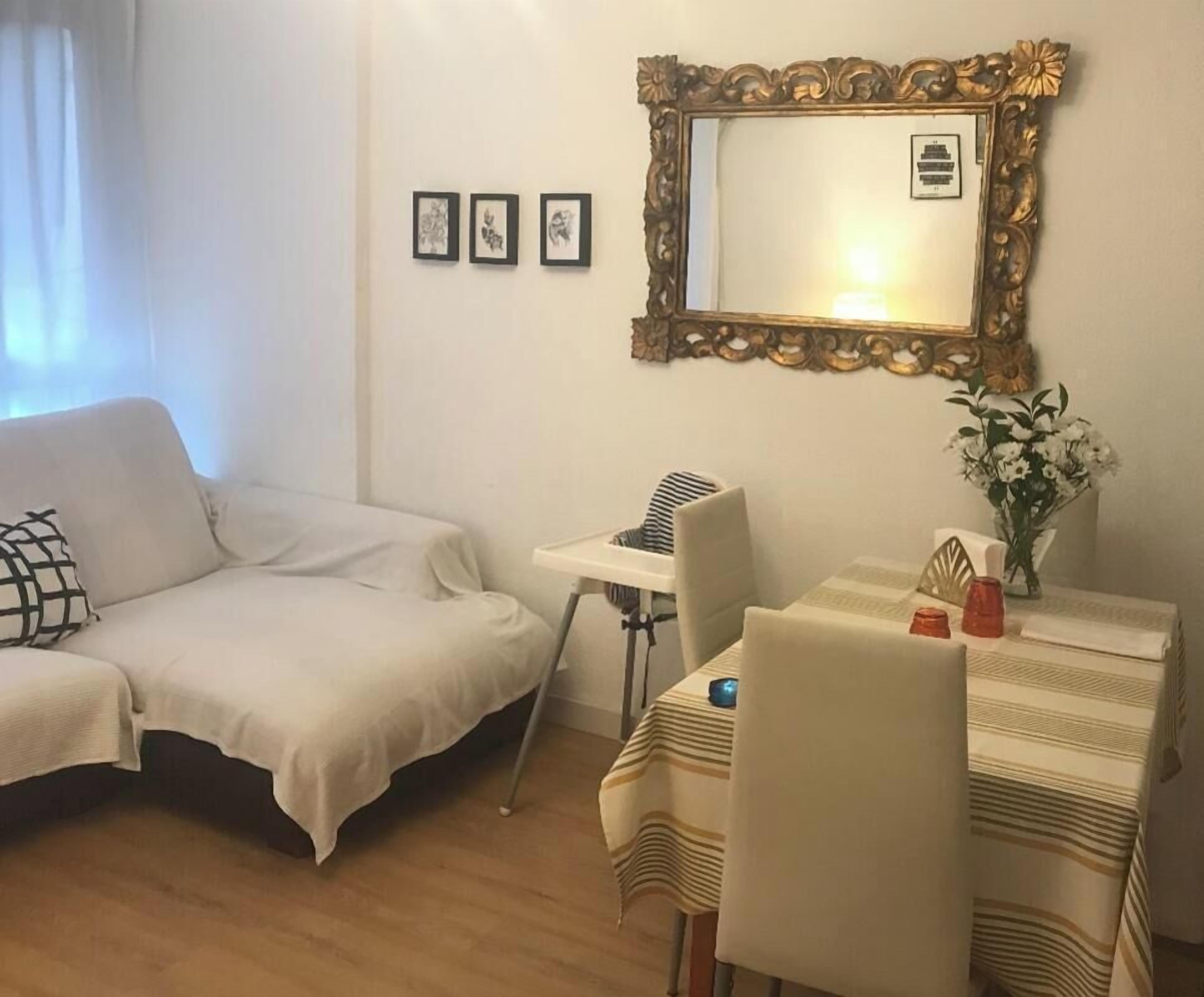 A room with a white sofa, a dining table with chairs, a small kitchenette, and a large mirror on the wall.