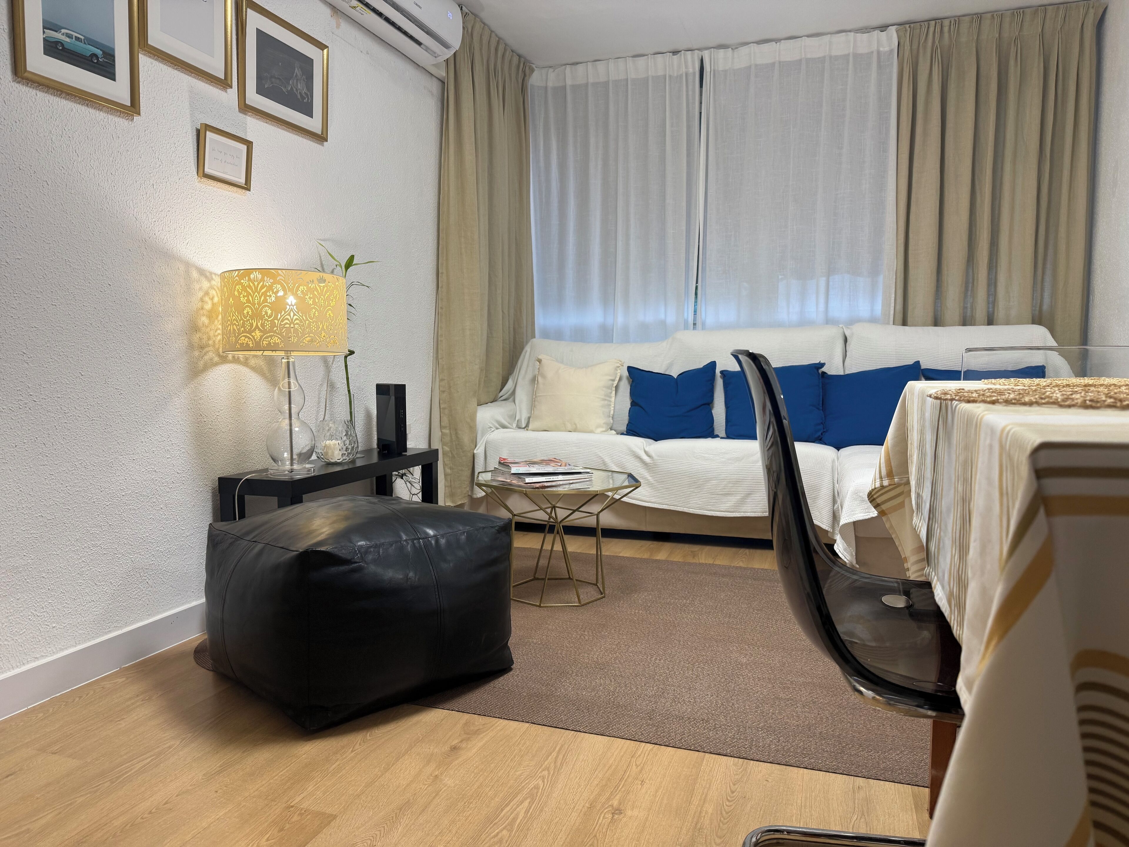A modern hotel room with a sofa, a black leather ottoman, a glass coffee table, and a lamp.