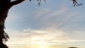 Cena de praia com pôr do sol, costa rochosa e montanhas ao longe.