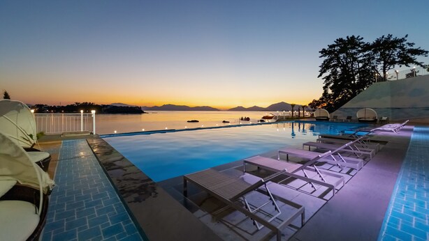 Piscina infinita com vista para o pôr do sol sobre a água, cadeiras de espreguiçadeira e um pequeno barco atracado nas proximidades.