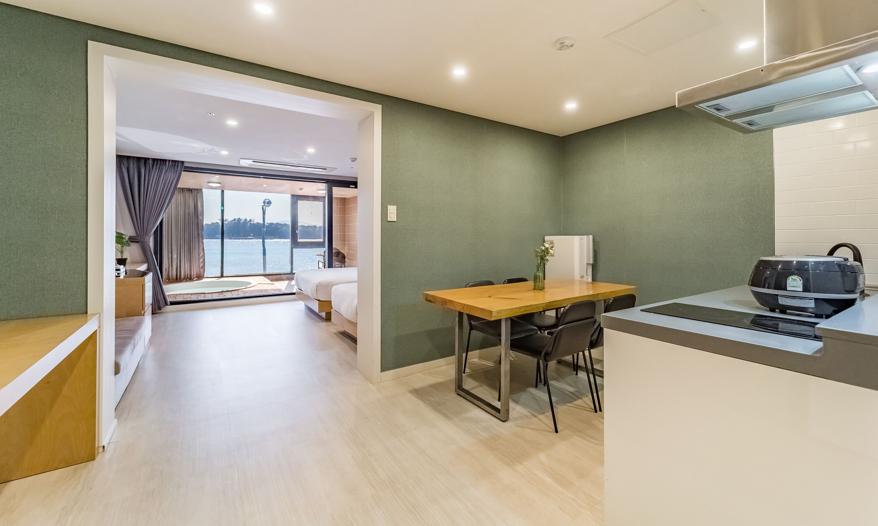 A modern hotel room with a wooden dining table, a small kitchen area, and a large window offering a view of the outdoors.