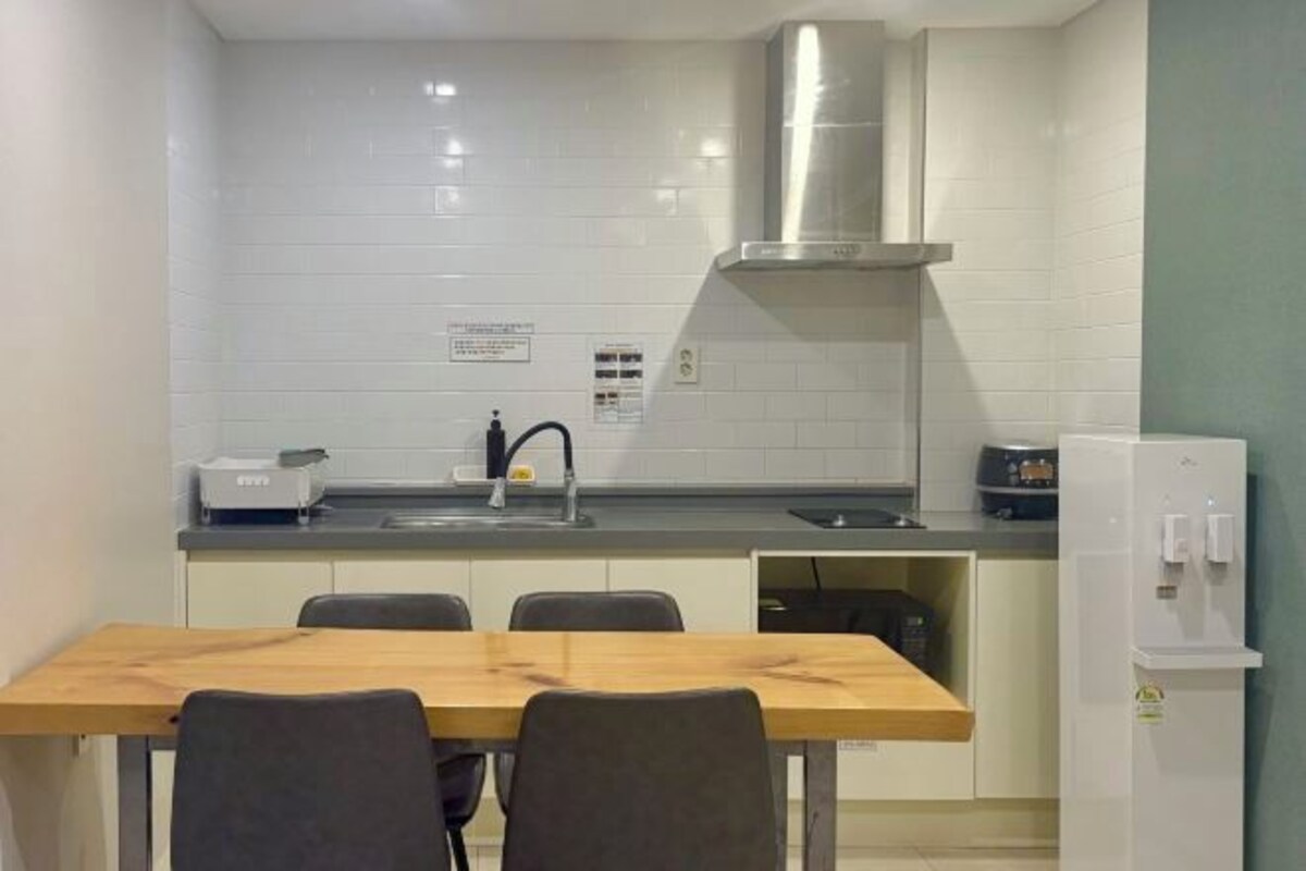 Una cucina compatta con piastrelle bianche, una cappa aspirante in acciaio inossidabile, un forno integrato, un frigorifero e un tavolo con sedie.