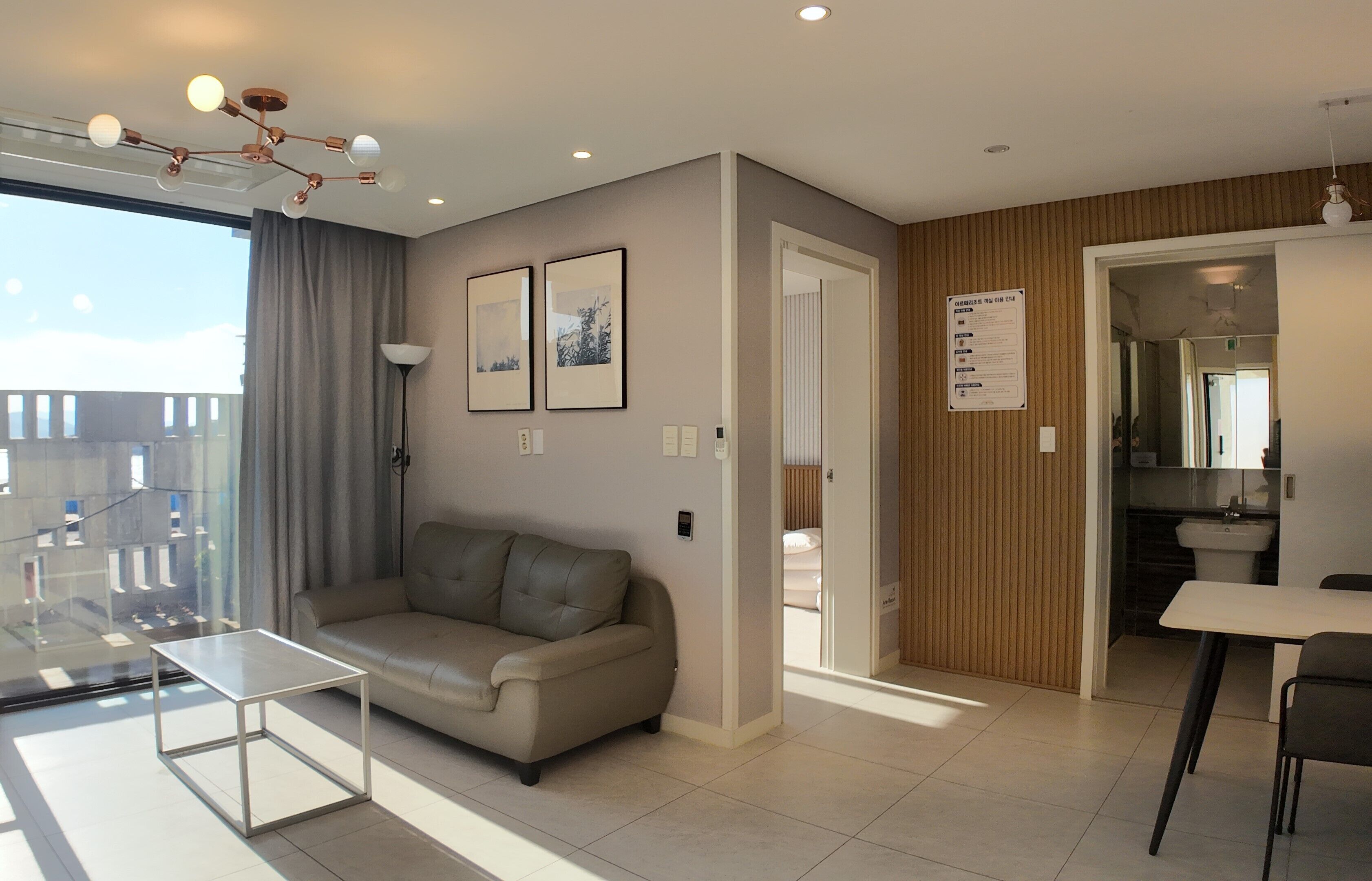A modern living room with a sofa, a glass coffee table, and a dining area with a table and chairs. There are framed pictures on the wall, a ceiling light fixture, and a view through a window.