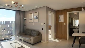 A modern living room with a sofa, a glass coffee table, and a dining area with a table and chairs. There are framed pictures on the wall, a ceiling light fixture, and a view through a window.
