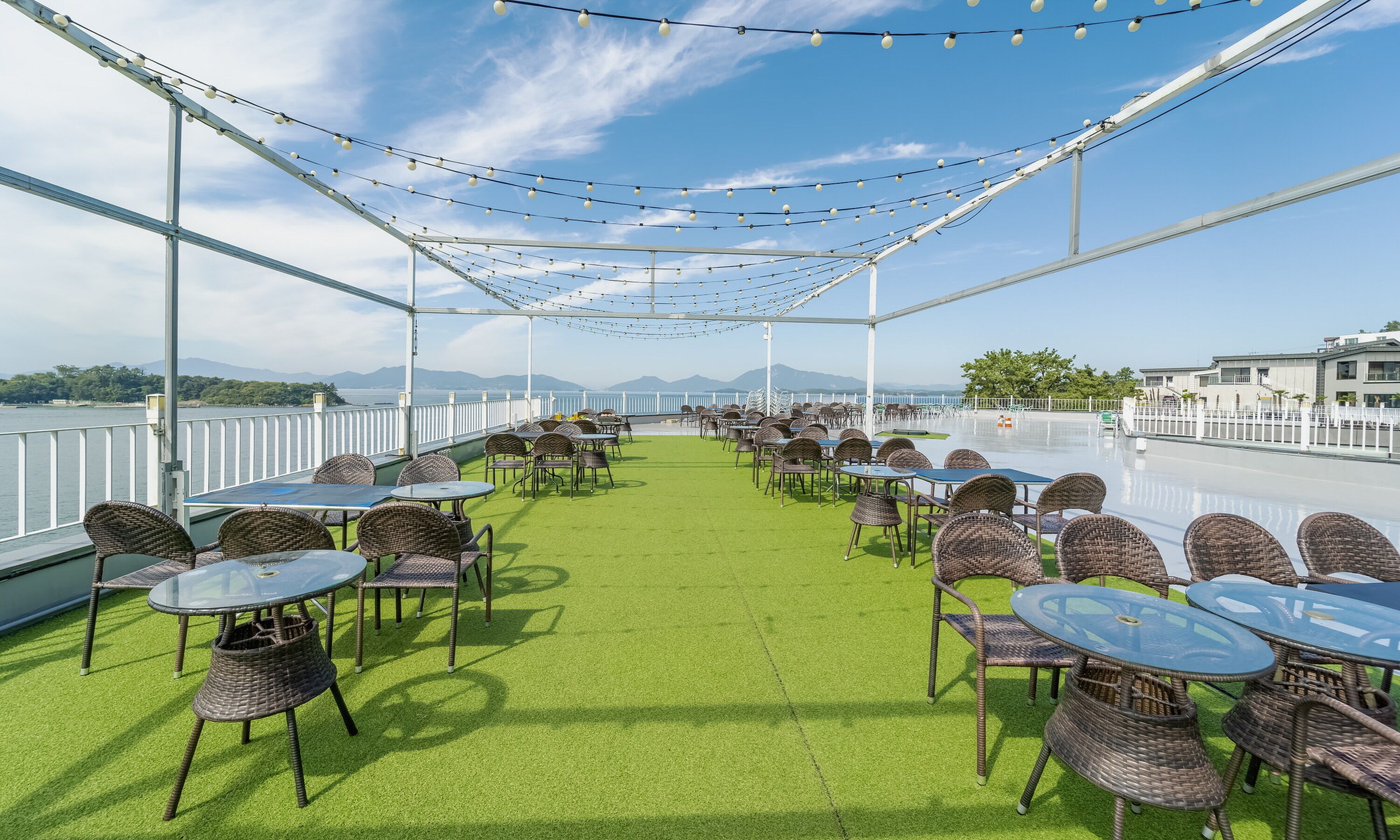 An outdoor terrace with tables and chairs, a view of the water, and a white fence.