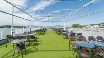 An outdoor terrace with tables and chairs, a view of the water, and a white fence.
