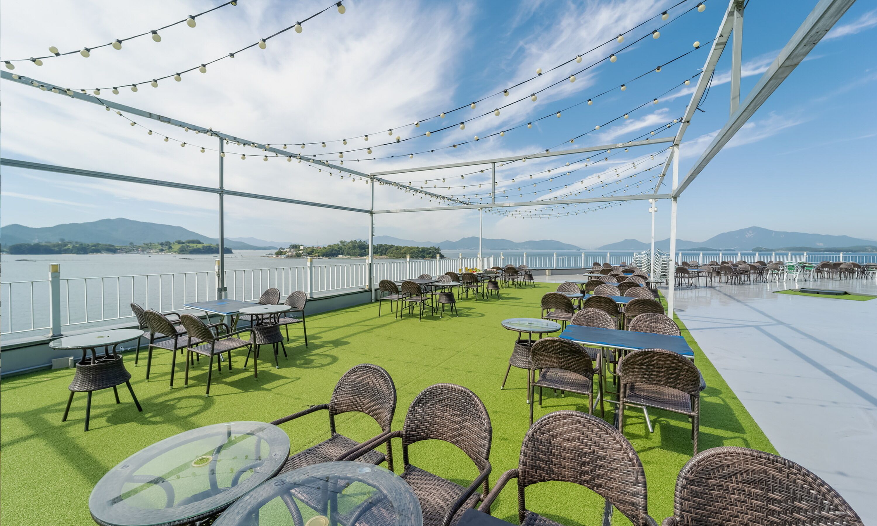 An outdoor seating area with tables and chairs, a view of the sea, and a white fence.