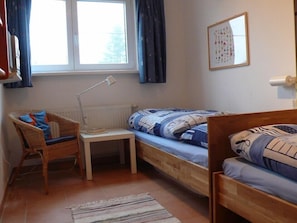 A small, cozy room with a wooden bed, a chair, a small table, and a window with curtains.