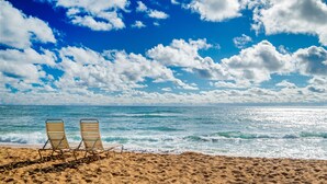 Ein Strand mit zwei leeren Liegestühlen, die auf den Ozean ausgerichtet sind, unter einem bewölkten Himmel.