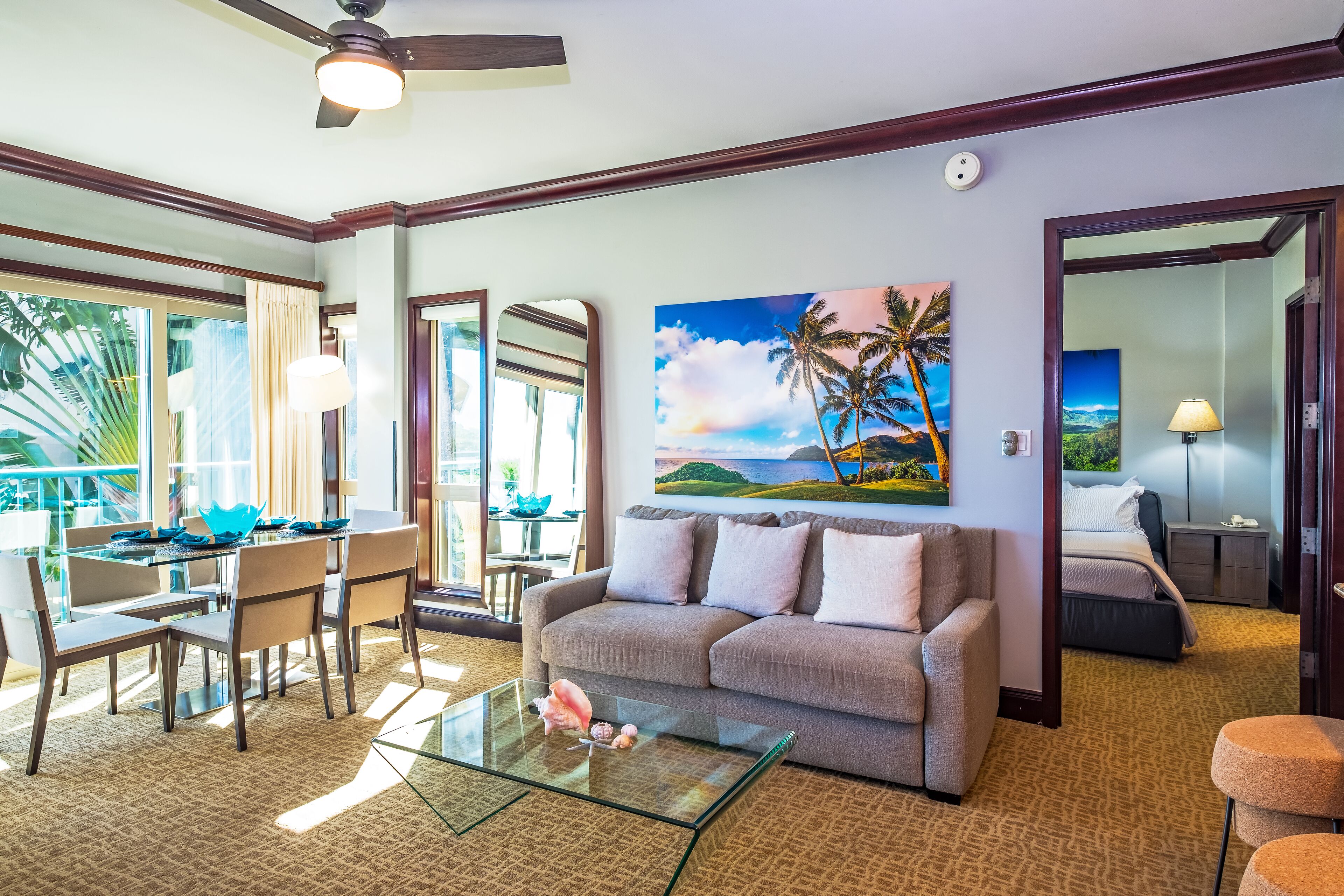Une chambre d’hôtel avec une table basse en verre, un canapé, un coin repas avec des chaises et un tableau représentant un paysage tropical.