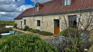 A two-story stone house with a red-tiled roof, a wooden door, and a small garden with a table and chairs.