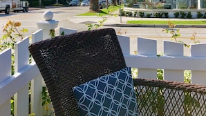 A balcony with wicker furniture and a white fence, overlooking a street with palm trees and a white building with a balcony. - Enjoy Ocean View from Large Private Patio, A/C & Garage (Corona del Mar)