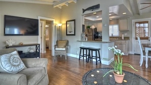 A spacious living room with a large flat-screen TV, a comfortable sofa, and a dining area with stools. - Enjoy Ocean View from Large Private Patio, A/C & Garage (Corona del Mar)
